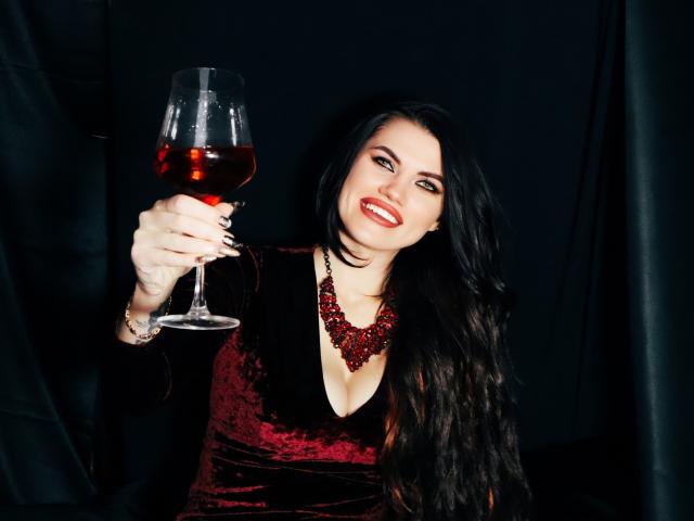MilaLoona, avec de longs cheveux noirs, une robe en velours rouge et un collier rouge, tenant un verre de vin rouge, souriante, exhibant ses seins. Fond noir..