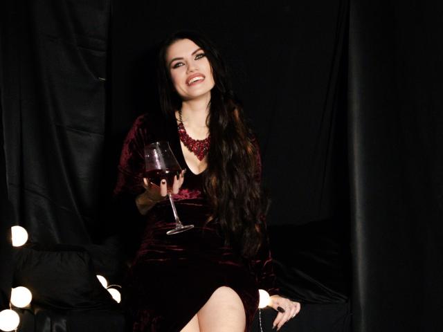 MilaLoona with long black hair, wearing a dark red velvet dress, holding a glass of red wine, smiling, sitting on a black chair, wearing a red necklace.