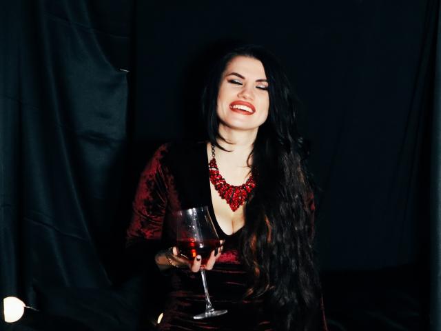 MilaLoona with long black hair, red velvet dress, and red necklace, holding a glass of red wine, smiling, showing off her tits, in a dark, moody setting.
