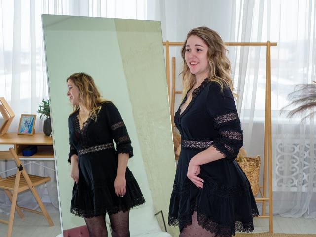 DoraSevi, capelli biondi e ondulati, vestito nero in pizzo, in piedi, sorridente, riflessa in uno specchio a figura intera, collant neri, mani sui fianchi, tende bianche, paravento in legno, pianta in vaso sullo sfondo..