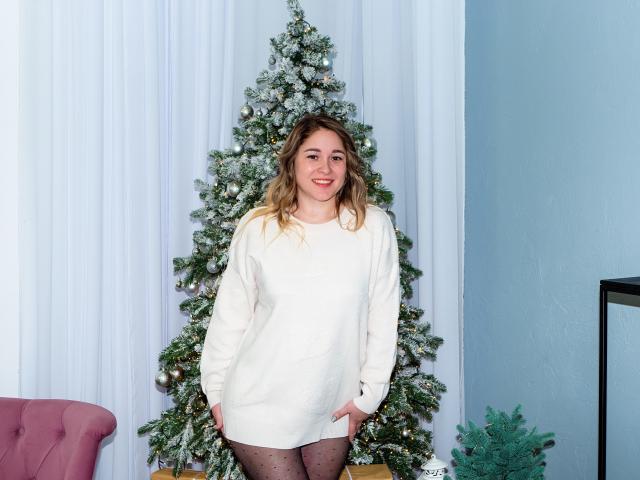 DoraSevi, in piedi di fronte a un albero di Natale decorato, indossa un maglione bianco, collant neri a pois, con le mani in tasca, sorridendo, capelli biondi, occhi azzurri, rossetto rosso..