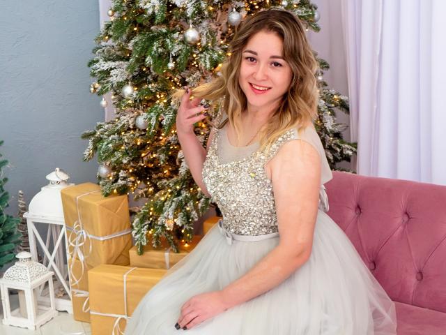 DoraSevi, con i capelli biondi, indossa un abito argentato scintillante e una gonna di tulle bianco, è seduta su un divano rosa, sorridendo e tenendosi i capelli, accanto a un albero di Natale decorato..