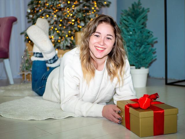DoraSevi, con i capelli biondi ondulati, sorridente, sdraiata su un tappeto bianco, indossa un maglione bianco, jeans blu, calze bianche, accanto a una scatola dorata con un nastro rosso, albero di Natale sullo sfondo..