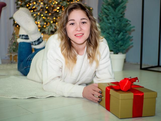 DoraSevi with blonde wavy hair, white sweater, and blue jeans, lying on floor, smiling, hands on gold gift box with red ribbon, white socks, Christmas tree in background.