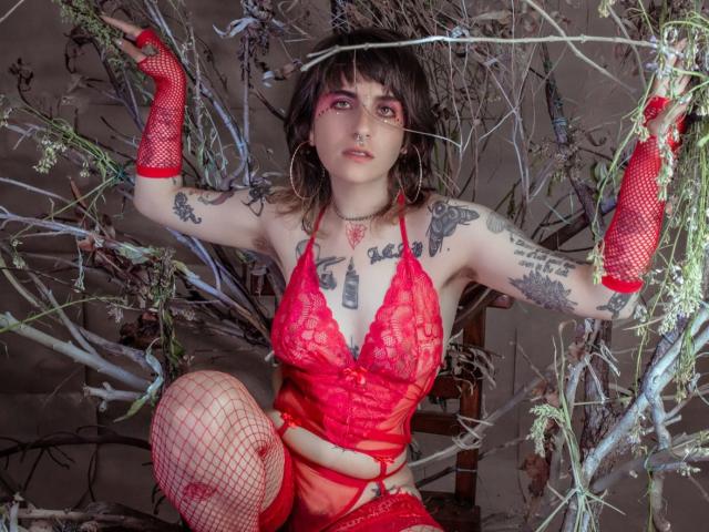 AmySpider, con tatuajes, lencería de encaje rojo, guantes de rejilla y medias altas rojas, está agachada entre las ramas, mostrando sus pechos, vulva y brazos, con aros en las orejas y maquillaje de ojos rosa..