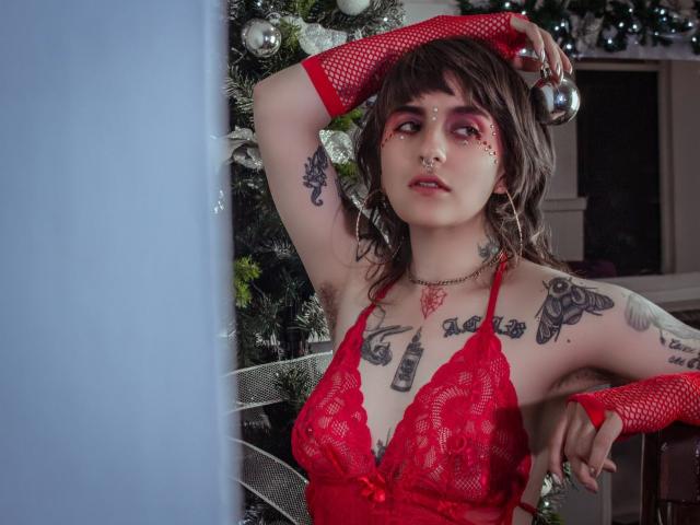 AmySpider with tattoos, red lace lingerie, fishnet arms, holding a ball, pink eye makeup, nose ring, and short brown hair, standing in front of a Christmas tree.