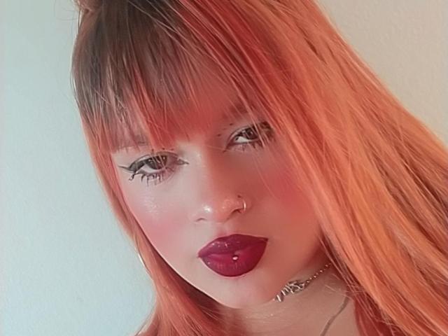 AmberMartinii has red-orange hair, dark eyeliner, purple lipstick, a nose ring, and a silver choker. Her pinkish skin and pouty lips are prominent. She looks straight at the camera.