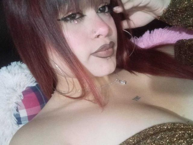 AmberMartinii with red hair, black eyeliner, and a nose piercing, showing her tits in a glittery top, lip ring, and necklace, lying down with a pink fluffy pillow.