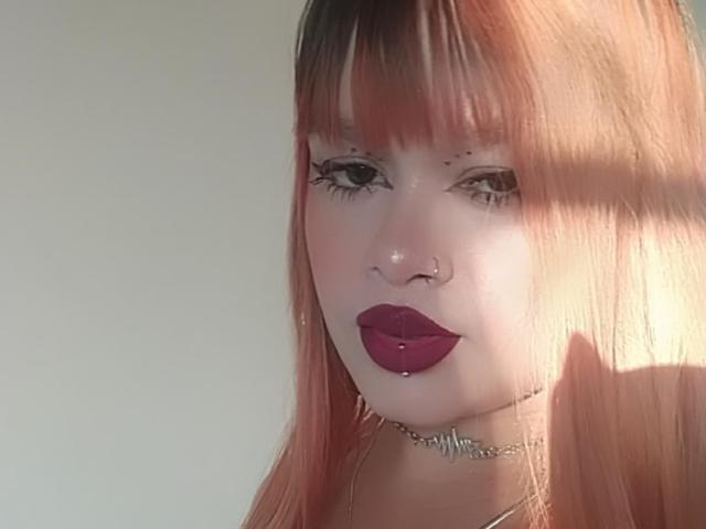 AmberMartinii, con la pelle chiara, i capelli rosa-arancio, la frangia, il rossetto viola scuro, l'eyeliner nero, un anello al naso e una collana con borchie, guarda leggermente a sinistra, illuminata da una luce soffusa..