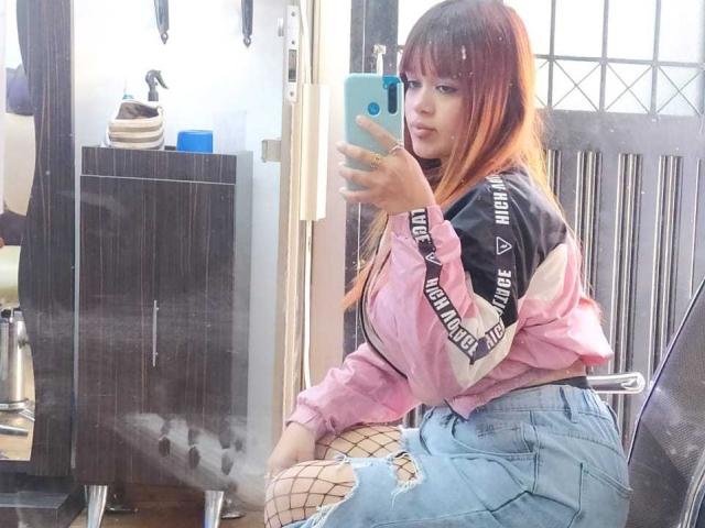 AmberMartinii mit rotbraunen Haaren, trägt ein übergroßes, pinkes und schwarzes Hemd, blaue Jeans und Fischernetzstrümpfe und macht ein Selfie vor einem Spiegel mit einem blauen Handy..