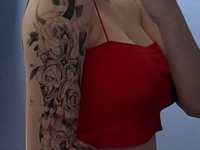 Destinis with a red spaghetti strap top, showing a detailed black and gray tattoo sleeve of roses on her right arm, and her small tits visible beneath the top.