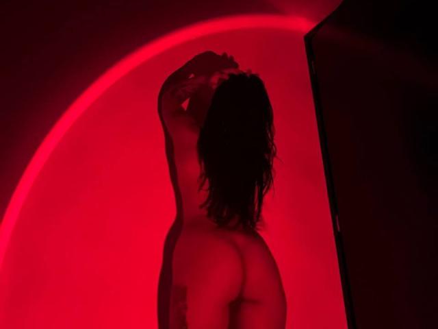 Amberv's naked back and ass are highlighted by red lighting, arms raised above head, wavy dark hair, standing in front of a dark screen. Red light creates a sensual glow.
