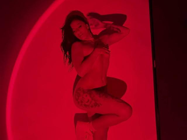 Amberv está desnuda, cubriéndose los pechos con la mano, en una habitación iluminada con luz roja, arrodillada con las nalgas levantadas, mostrando un tatuaje en el muslo y llevando tacones blancos..