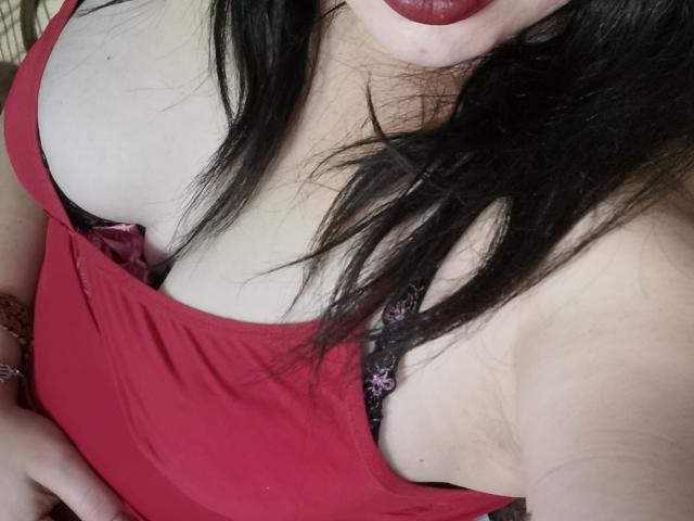 El vestido rojo de KaraaGul revela un sujetador de encaje negro, mostrando sus senos y un poco de escote. Tiene el pelo oscuro, lápiz labial rojo y un tono de piel claro..