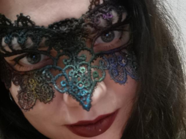 Il viso di KaraaGul, con i suoi capelli scuri e ondulati, indossa una mascherina di pizzo colorata che le copre gli occhi, e ha un rossetto rosso intenso. I suoi occhi sono verdi, e la mascherina presenta motivi elaborati..