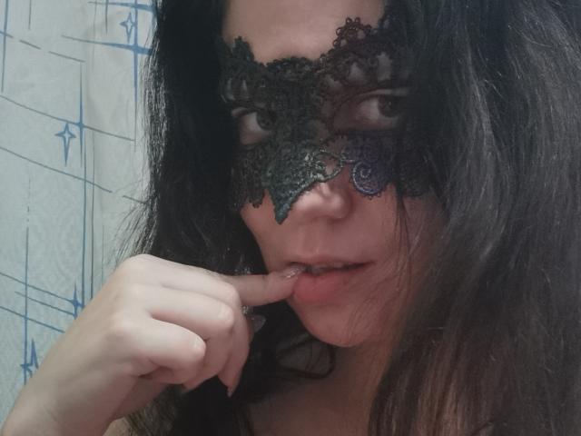 KaraaGul, com cabelos escuros e ondulados, usando uma máscara de renda preta, mordendo o dedo, exibindo um sorriso provocador e sedutor, seus olhos parcialmente escondidos pela máscara elaborada..
