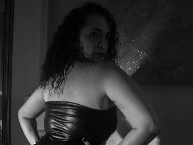 Scarlett Vega, con cabello oscuro y rizado, mirando por encima del hombro, vestida con una blusa negra de cuero sin tirantes, con los brazos ligeramente levantados, mostrando su espalda y trasero desnudos. Blanco y negro..