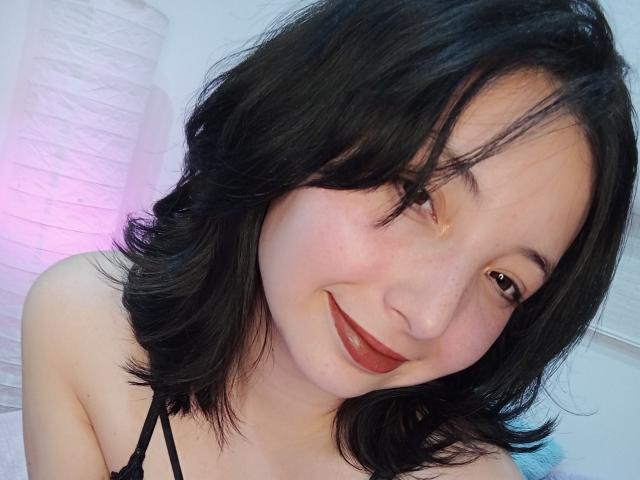 BarbaraJonesX mit schwarzen, schulterlangen Haaren, lächelnd mit rotem Lippenstift, trägt ein schwarzes Top mit Halter, hat helle Haut, braune Augen und leicht gewelltes Haar, in einem ungezwungenen Selfie..
