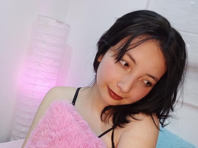 BarbaraJonesX with black hair, fair skin, and red lipstick, smiling while holding a pink fluffy pillow, wearing a black spaghetti strap top, standing next to a pink lamp.