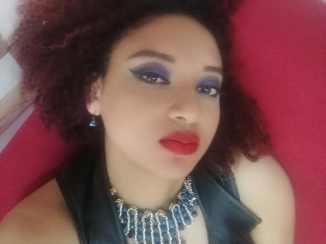 VioletaSwitch has curly red hair, purple eyeshadow, red lipstick, blue earrings, and a black necklace with blue gemstones. She's wearing a black sleeveless top.
