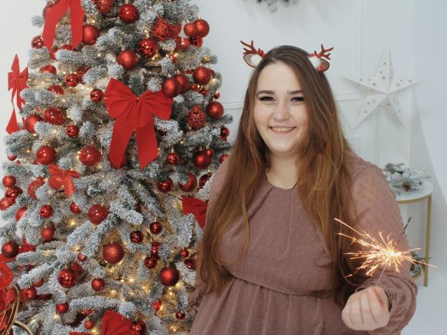 DaniellaGraceZZ with red reindeer antlers, holding a sparkler, wearing a brown dress, smiling next to a decorated Christmas tree with red ornaments and red bows. Boobs visible under dress.