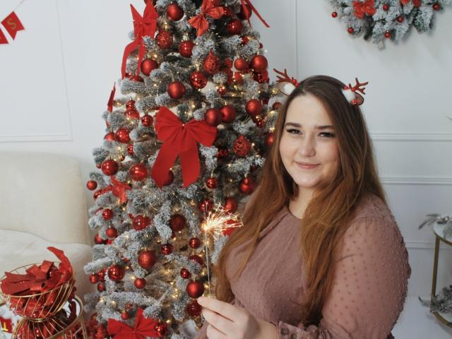 DaniellaGraceZZ with long brown hair, wearing a pink, sheer top, holding a sparkler, standing next to a red and silver Christmas tree with red bows and ornaments.
