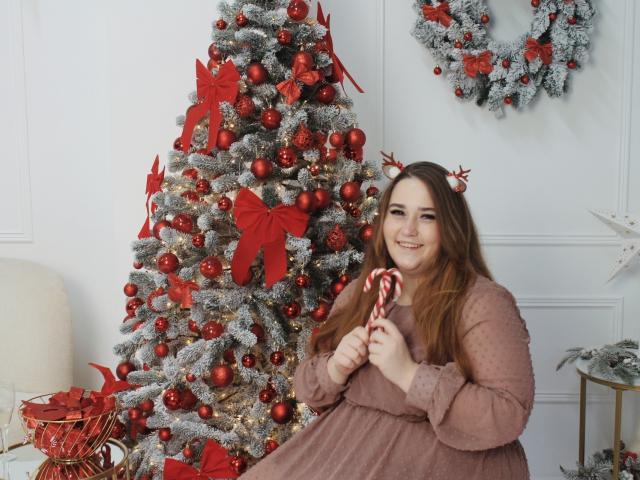 DaniellaGraceZZ with long brown hair, wearing a pink dress, holding a candy cane, smiling next to a red and silver Christmas tree with red ornaments and red bows.
