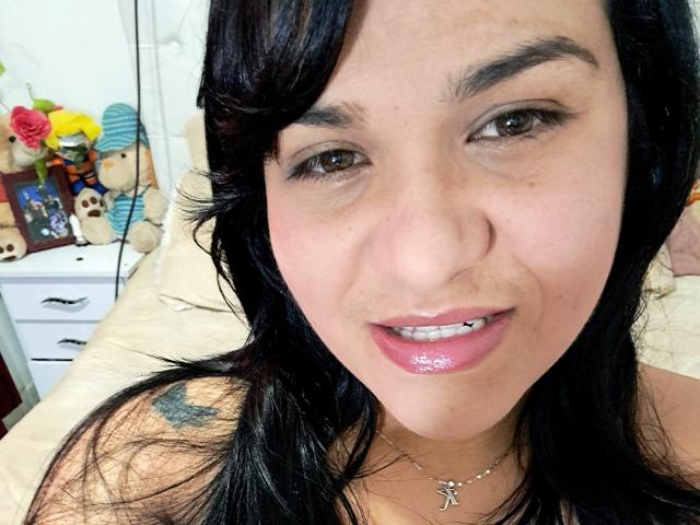 VivianaThompson has black hair, brown eyes, and glossy pink lips. She's smiling with a small tooth gap, wearing a necklace, and has a tattoo on her right shoulder.
