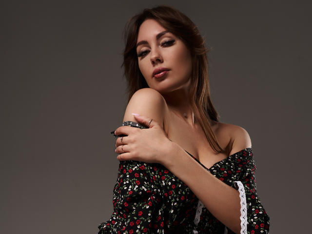 NinaTender with her tits slightly exposed, wearing a floral off-shoulder dress, arms crossed over her chest, brown hair, subtle makeup, gray background, soft expression.