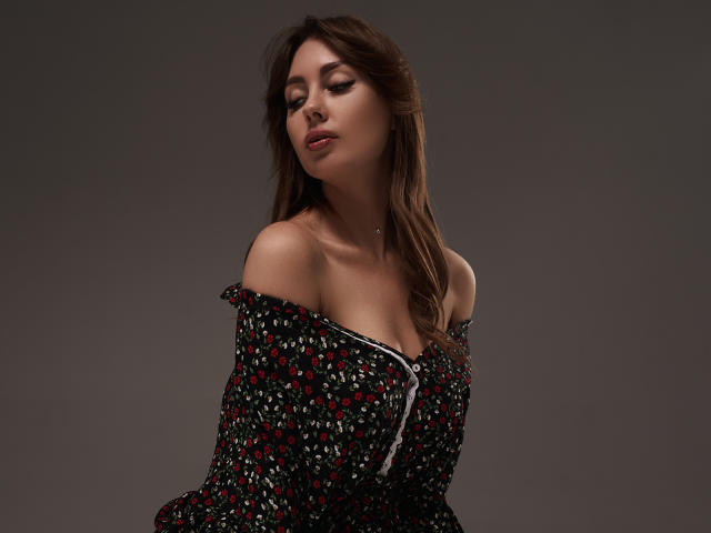 NinaTender has her eyes closed, wearing a black off-shoulder dress with red and white floral patterns, showing her tits and a slight cleavage, with brown wavy hair.