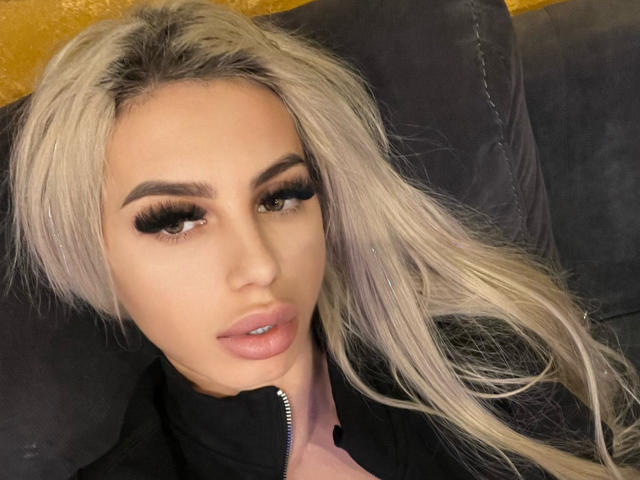 LisaaKiraa, avec de longs cheveux blonds platine, des sourcils épais, des cils longs et des lèvres roses brillantes, portant un haut noir, assise sur un canapé gris foncé..
