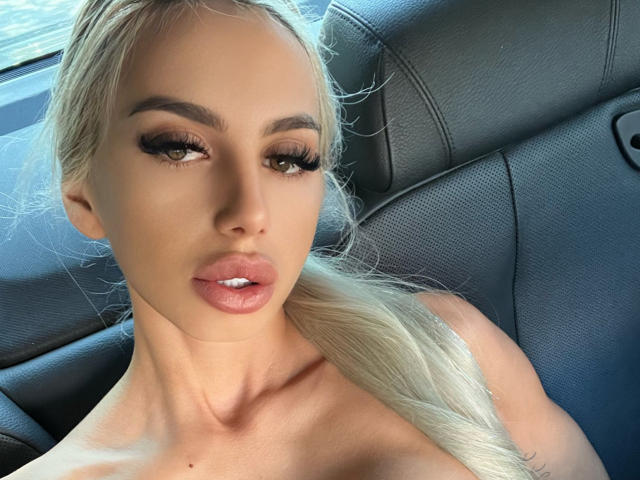 LisaaKiraa with blonde hair, heavy eyelashes, and glossy pink lips, looking at the camera with a slight pout, sitting in a car with black leather seats.