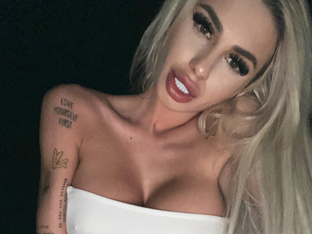LisaaKiraa with blonde hair, full lips, and large tits, wearing a white strapless top, showing tattoos on her shoulder and upper arm, against a dark background.