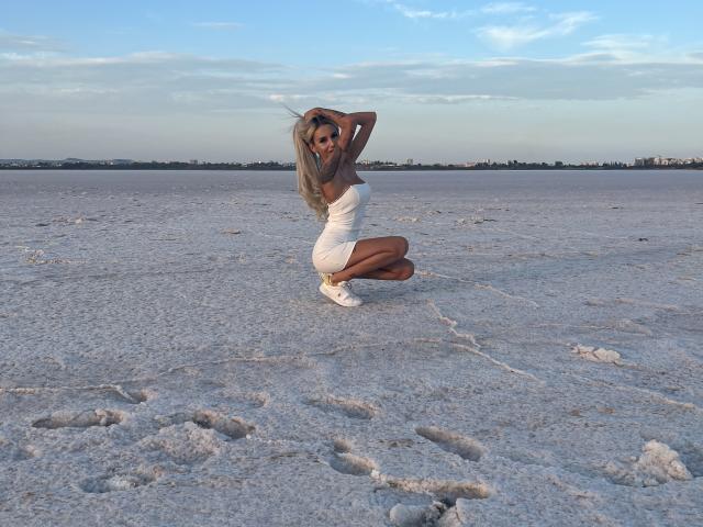 LisaaKiraa, hurkend op zoutvlaktes, draagt een grijze top en witte shorts, haar handen in haar blonde haar, bruine huid, witte sneakers, waarbij haar achterkant zichtbaar is, onder een heldere lucht..