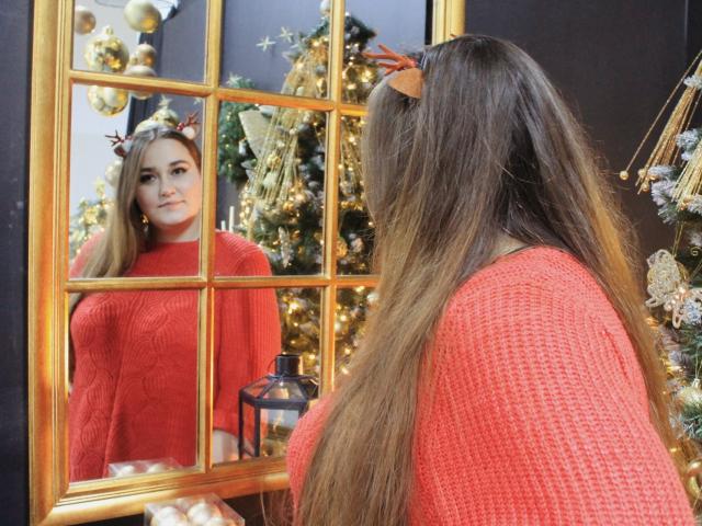 DaniellaGraceZZ with long brown hair, red sweater, and antler headband, staring at her reflection in a gold-framed mirror, showing her small tits, in a festive, decorated room.