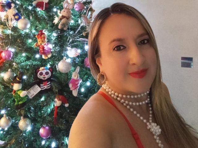 Charlize, con capelli biondi, vestito rosso, collana di perle e rossetto rosso, sorride accanto a un albero di Natale decorato con ornamenti colorati e luci. Il seno è visibile nel vestito rosso..