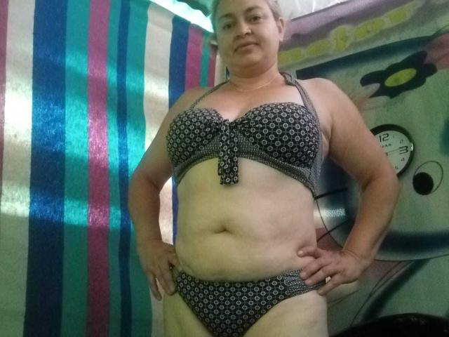 SalomeAriza se tient dans une pièce, vêtue d'un soutien-gorge et d'une culotte à motifs noirs et blancs, exposant ses seins volumineux et tombants ainsi que son ventre, les mains sur les hanches, avec une expression neutre..