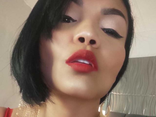 ExoticLyla with black bob, red lipstick, and thick black eyeliner, slightly parted red lips showing white teeth, wearing gold earrings, close-up shot.