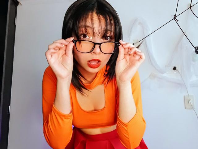 BarbaraJonesX with black glasses, orange crop top, and red skirt, holding glasses, looking surprised, with red lipstick, medium tits, and straight black hair.