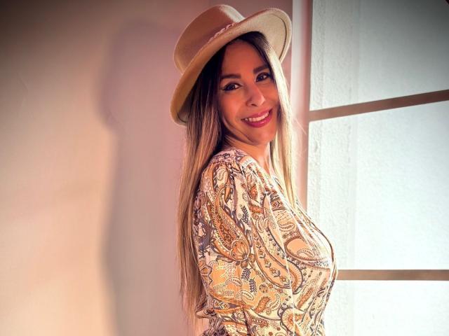 VictoriaJhonnes with long blonde hair, smiling, wearing a beige hat and a patterned shirt, showing her tits in a bright room. Her ass is not visible.