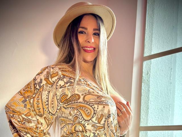 VictoriaJhonnes, con lunghi capelli biondi, indossa un cappello beige e una camicia con motivo paisley, sorride con il rossetto rosso, mostrando il seno, ed è in piedi vicino a una finestra..