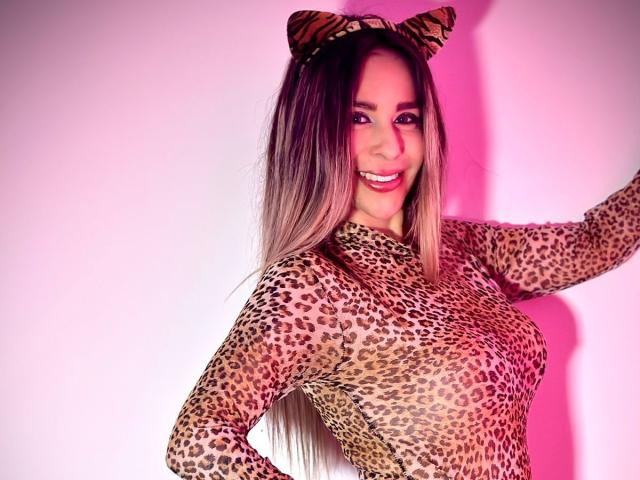 VictoriaJhonnes with long blonde hair, wearing a leopard print top and cat ear headband, smiling, showing her tits, in a pink-lit shot.