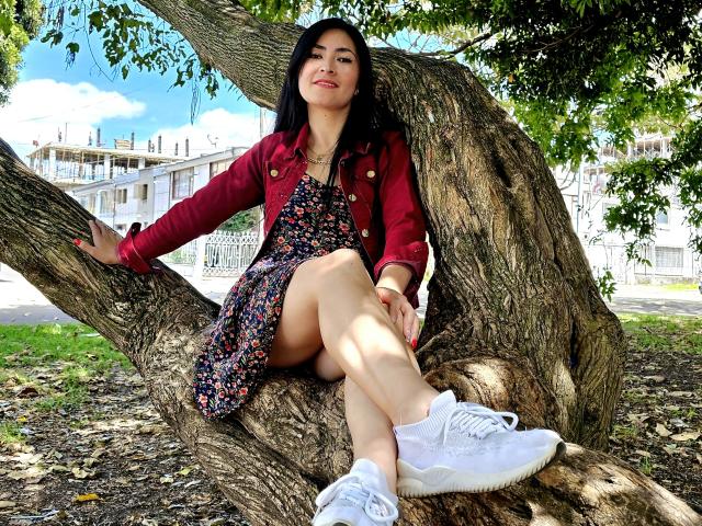 JainaRogers, con capelli neri, giacca rossa, vestito floreale e scarpe da ginnastica bianche, seduta su un ramo spesso di un albero, gambe incrociate, sedere leggermente sollevato, sorridente, in un parco soleggiato..