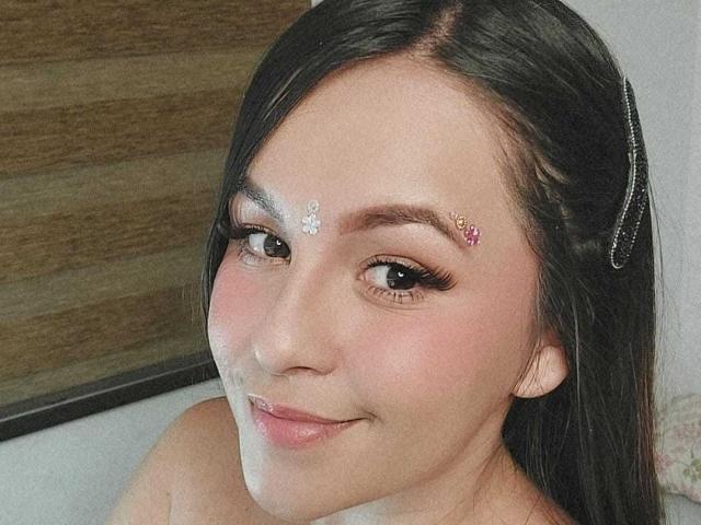 YudyPatty ha i capelli castano scuro, un piccolo fiore bianco e una gemma rosa sulla fronte, con lunghe ciglia e un sorriso carino. Sorride leggermente, mostrando le sue labbra rosa..