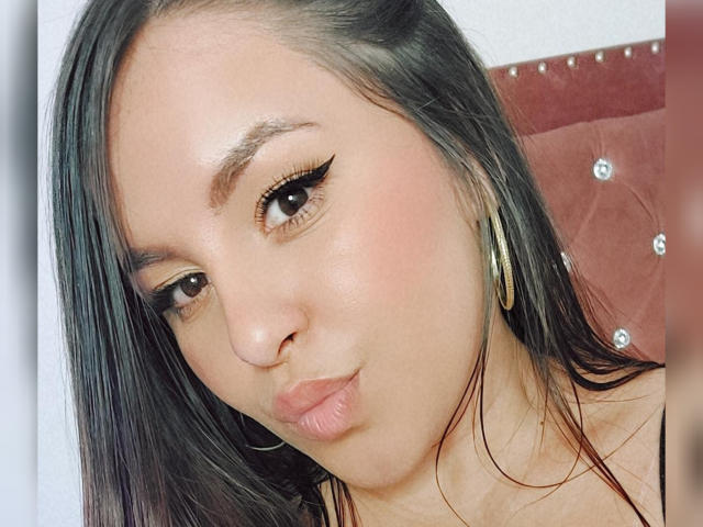 YudyPatty, avec ses lèvres pulpeuses, ses cheveux noirs et lisses, et son trait d'eye-liner noir épais, portant des boucles d'oreilles dorées, regardant l'appareil photo avec un léger froncement de sourcils..