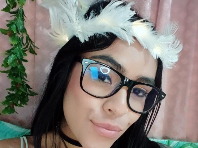 StellaBeckker with black glasses, white feathered headpiece, and pinkish-brown skin, wearing a black choker, smiling at the camera, green leaf hanging on the left.