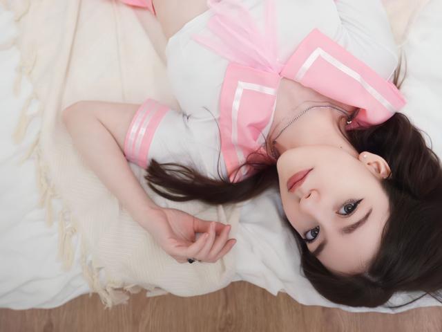 VinnaSkyHott, allongée sur un lit, portant une tenue de marin rose et blanche, regarde vers le haut avec ses yeux marron, ses lèvres roses et ses cheveux brun foncé, montrant une main délicate et un collier..