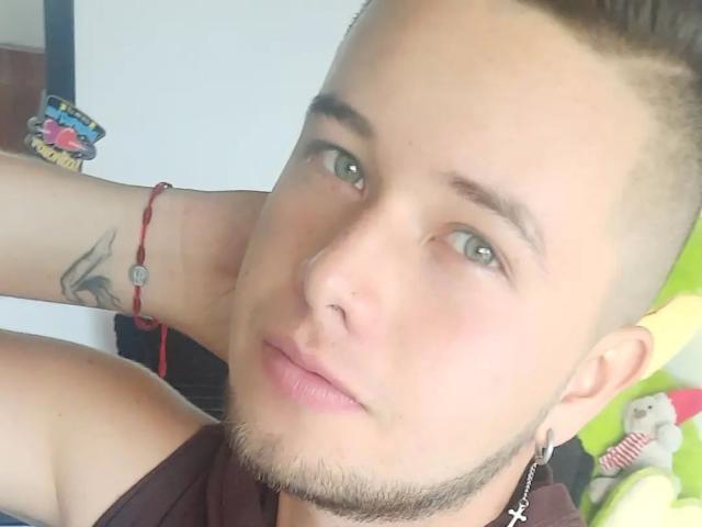 CuteManX mit kurzen, braunen Haaren, grünen Augen und einem Nasenring, trägt ein braunes Hemd, hat ein Tattoo am Arm und ein rotes Armband. Sein Gesicht ist leicht schmollend..