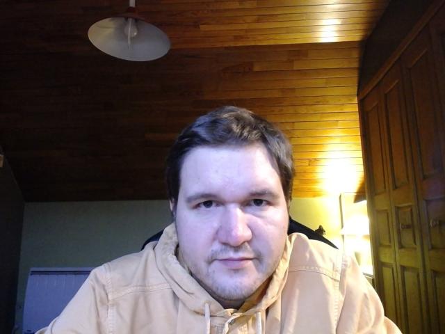 NectarineFad has a chubby face, short brown hair, a light beard, and wears a beige hoodie with a black backpack strap over his shoulder. Ceiling light is visible.