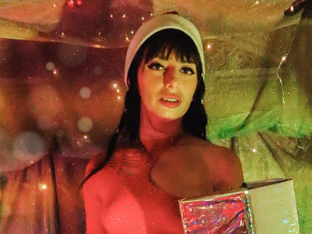 LeticiaFlorenz mit dunklem Haar, die eine weiße Mütze trägt, oben ohne, hält eine glitzernde Schachtel, rotes Licht, funkelnder Hintergrund, blickt in die Kamera mit leicht geöffnetem Mund und zeigt ein kleines Tattoo..